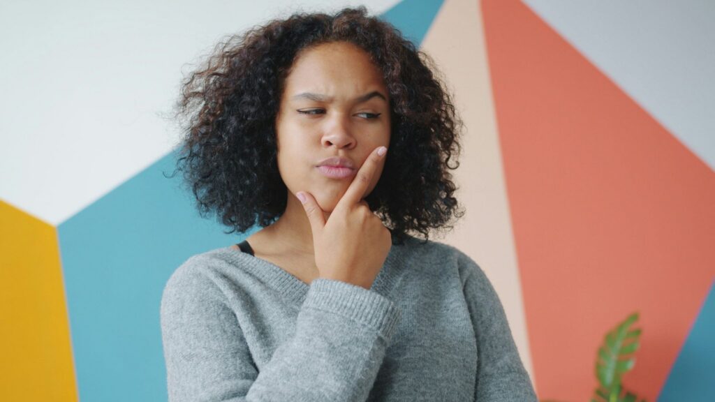 A woman with dark curly hair and mid-brown skin looks quizzically to the right, holding her chin between forefinger and thumb. The image suggests skepticism.